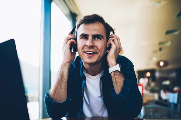 Handsome smiling creative male blogger spending leisure time indoors and listening to favorite radio station online via headphones.Cheerful hipster guy enjoying recreation with music and good mood