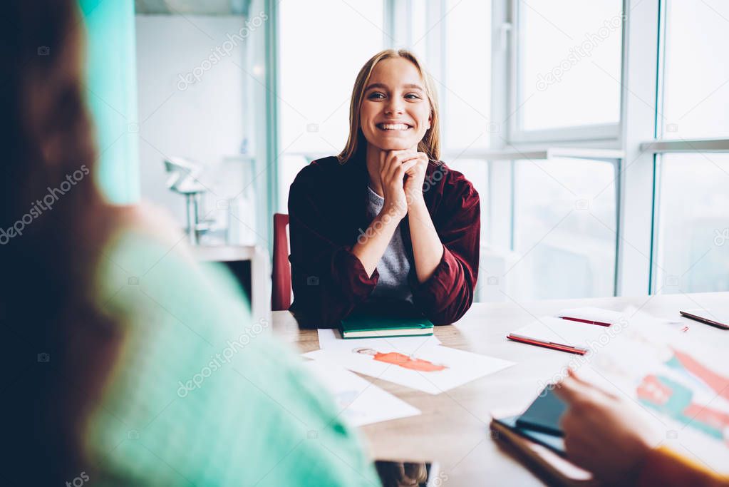 Cheerful hipster girl with blonde hair laughing during work break sitting at meeting table with sketch of design clothes.Happy student discussing successful ideas for developing own business in office