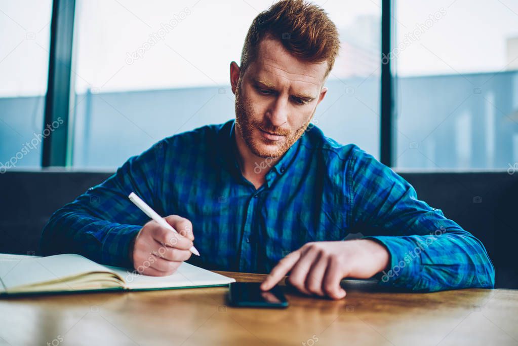 Skilled student with red hair writing down text information from website on smartphone sitting in university interior.Smart hipster guy making notes for coursework reading text from cellular