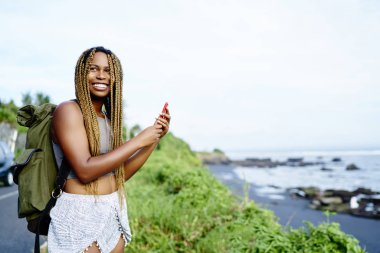 Mutlu afro Amerikalı genç kadın Smartphone'da tropikal ortamda resimlerini yaparken gülüyor sırt çantası ile. Pozitif kadın turist koyu tenli hücresel adada seyahat fotoğraf çekmek