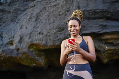 Neşeli karanlık kadın jogger mutlu serin müzik radyo için sabah idmanları sahil şeridi üzerinde derili. Pozitif genç kadın oyuncu cep eğitim önce bağlı kulaklık üzerinden ses dinleme