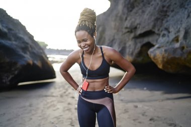 Sabah egzersiz sırasında bir şekilde gülerek eraphones komik ses şarkıları dinleme şık spor giyim giymiş genç kadın neşeli koyu derili. Afro Amerikan kadın eğitim üzerinde müzik keyfi deniz kıyısı
