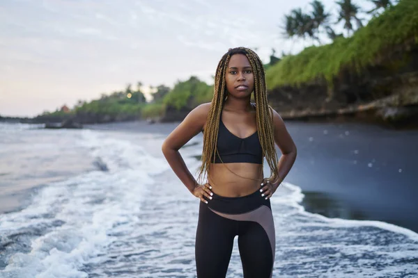 Koyu tenli ve okyanus ile spor sabah eğitimden sonra yıkanmış kıyısında duran güzel dreads ile motive genç kadın portresi. Kameraya bakarak eşofman giymiş Afro Amerikan kadın jogging yapan