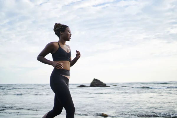 Side view of afro american female jogger dressed in stylish sport clothes running along coastline washed by ocean water.Sportive woman with dark skin doing morning cardio training on seashore