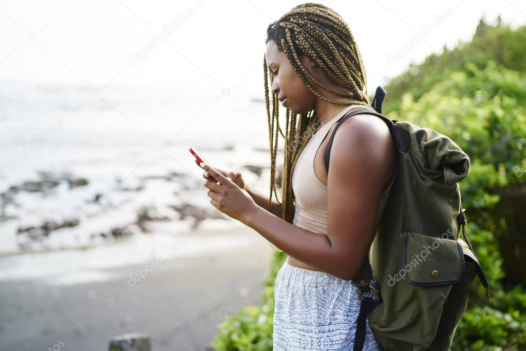 Dark skinned young tourist with dreads searching online navigator on smartphone standing outdoors.Afro american traveller with backpack sending sms on cellular strolling in tropical locality