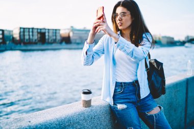 Yoğun hippi kız smartphone uygulama, eğlence sırasında takipçileri cep telefonu üzerinden paylaşmak için resim yapma genç kadın blogger kullanarak Nehri köprüsü üzerinde oturan rahat kıyafet alma fotoğraf