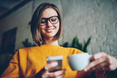 Şık saç kesimi ve süre komik fotoğraf uygulama yükleme ve smartphone ücretsiz Wi-Fi bağlantısıyla Cafe takipçileri ile mesajlaşma kapuçino keyfi gözlük ile pozitif kadın internet kullanıcı