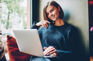 Coworking ücretsiz Wi-Fi kullanarak kendi web sitesi geliştirmek için yararlı bilgi aramak için modern dizüstü bilgisayar ile pencere üzerinde dinlenme sırasında kameraya gülümseyen güzel hippi kız portresi