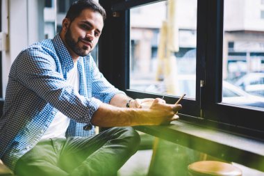 Sakallı yakışıklı kamera kamera, genç erkek öğrenci bloglama ağlar kahve molası sırasında telefonda portresi bakarak smartphone üzerinden sohbet iletileri gönderme kafede oturan bakarak