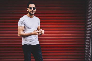 Sohbet için smartphone kullanarak şık giyimli erkek portresi yarım uzunlukta online promosyon arka planda, hücresel açık havada üzerinden mesaj gönderme kamera bakarak yakışıklı hipster adam ayakta