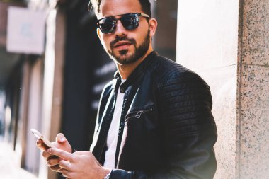 Boncuklu erkek güneş gözlüğü şehir sokak, hipster adam açık havada modern telefon kullanarak sosyal ağlarda sohbet smartphone ayakta aracılığıyla metin iletileri gönderirken kameraya bakarak Resim kırpılmış