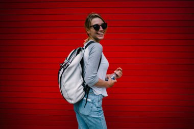 Camer seyir trendy sırt çantası ile poz güneş gözlüklü kadın turist gülümseyen neşeli hippi kız rahat kıyafet ayakta parlak kırmızı duvar açılış su şişesini, yarı uzunluk portre