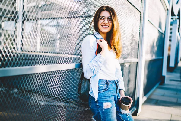 开朗的年轻女子在休闲服装看着站在城市设置背景 微笑时髦的女孩在时尚的眼镜享受自由时间举行咖啡去户外休息 — 图库照片