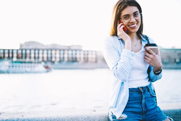 欢快的时髦女孩的肖像与咖啡去谈手机站在复制空间附近的广告户外 微笑的女性旅行者使用漫游拨打国际电话 — 图库照片