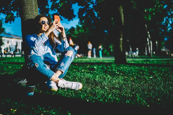座って公園の木の下で十代の内気な少女のイヤホンで音楽を聴くと Selfie 草で春の日の間に休憩を作るスマート フォンのカメラで写真を撮ってサングラスの若い女性を笑顔 — ストック写真