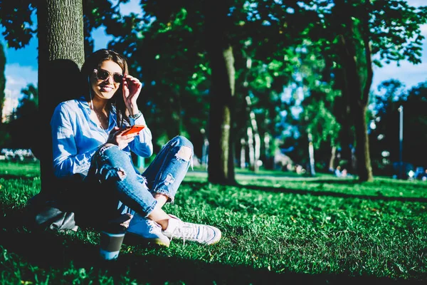 電話でリスニングのプレイリストの曲を再現する陽気な若い女性の肖像画を携帯とイヤホン アプリケーションを使用して音楽を楽しんでいる公園の芝生の上に座っているサングラスに笑みを浮かべて内気な少女 — ストック写真