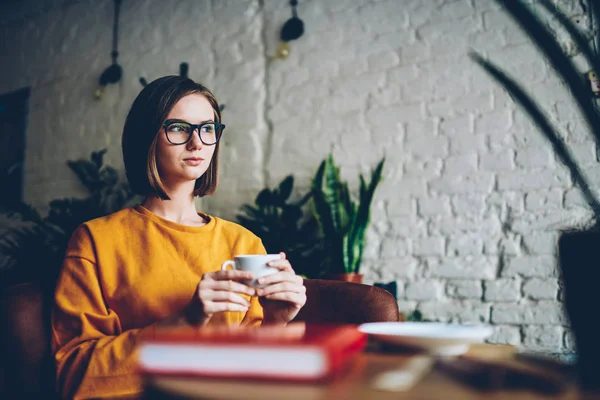 Smart young woman with short haircuts and eyeglasses pondering on new design startup project with cup of tasty coffee resting in cafeteria interior.Serious female thinking on business idea