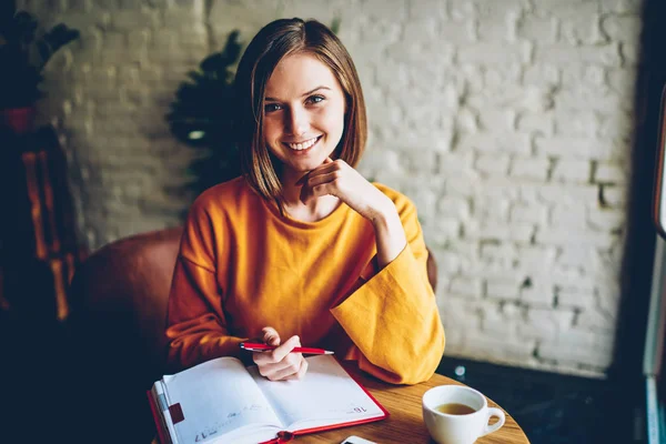 Half length portrait of beautiful cheerful student with stylish short haircut smiling at camera making notes in notebook at cup of coffee in morning time.Positive female owner writing working plan