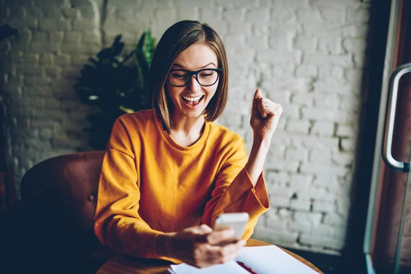 Emotional female in eyewear happy from getting good job offer via mail on digital cellular.Joyful hipster girl holding smartphone celebrating victory in online competition resting in stylish cafeteria