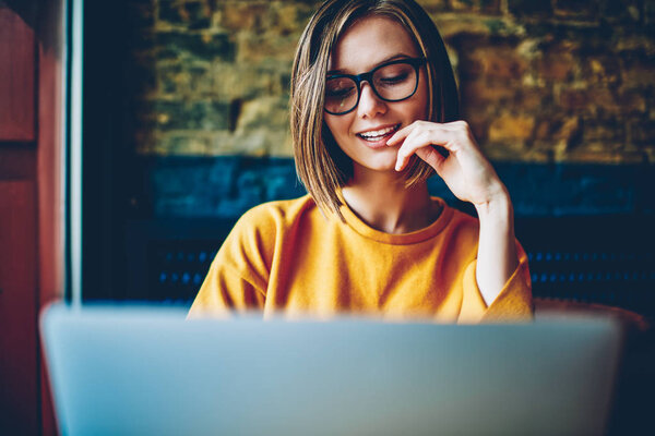 Cheerful attractive female student with eyeglasses and short haircut chatting online in social networks with friend on modern computer using free high speed internet connection sitting in coffee shop