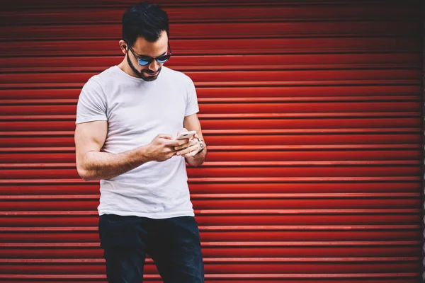 Handsome hipster guy banking via application on mobile phone standing outdoors on promotional background,young trendy dressed man text messages on smartphone chatting in social networks with followers