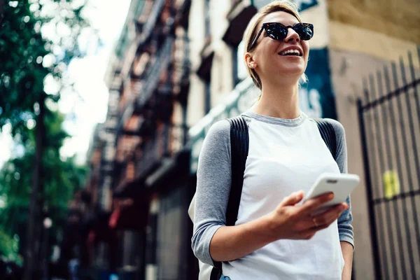 都心の移動に ネットワークで雑談する持株スマート フォンとの散歩を楽しんでいる肯定的な若い女性の携帯電話アプリケーションを使用して散歩のサングラスの笑みを浮かべて内気な少女 — ストック写真