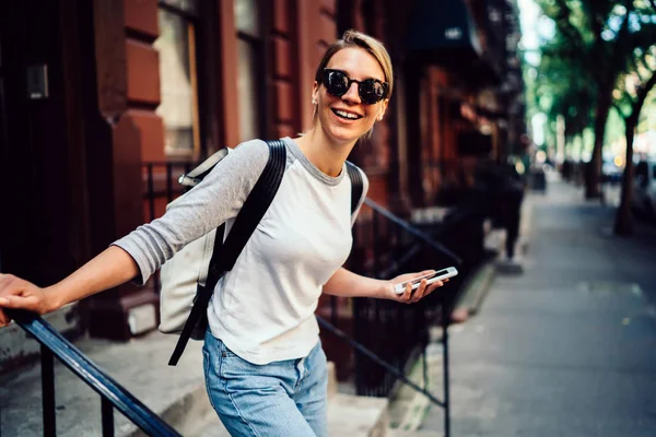 半长的开朗年轻女子的肖像离开大厦笑着拿着电话 微笑着的时髦女孩在太阳镜下周末积极愉快地漫步在市中心与 Smartphon — 图库照片