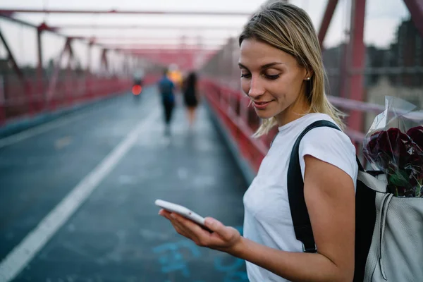 若い女性のウェブサイトや橋を散歩しながら携帯電話経由での情報の読み取りを参照する内気な少女と花 バックパックを運ぶ都市設定に歩きながらスマート フォンでメールをチェック — ストック写真