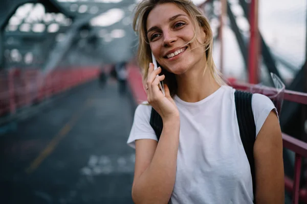 Cheerful hipster girl satisfied with operator consultancy talking on mobile phone while walking on urban setting, smiling young woman having telephone conversation in roaming strolling on bridg