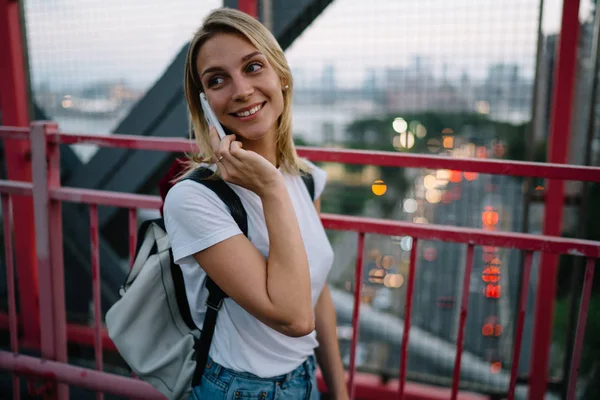 Smiling hipster girl smiling during mobile phone conversation with friend strolling with rucksack,positive female traveler having telephone conversation using cheap tariffs in roaming while walkin