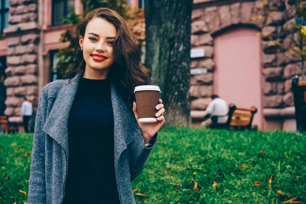 赤い唇屋外で立って カメラ目線と笑顔プロモーションのカラフルな背景の中 おいしいコーヒーを手で押しと陽気な流行に敏感な女の子の半分の長さの肖像画 — ストック写真
