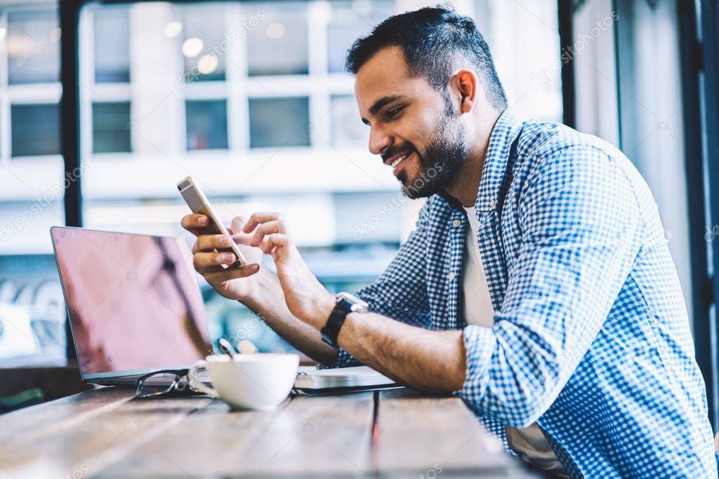 Cheerful male person text messages on smartphone chatting in social networks during coffee break,smiling young man checking mail on cellular pleased with getting discount for mobile internet