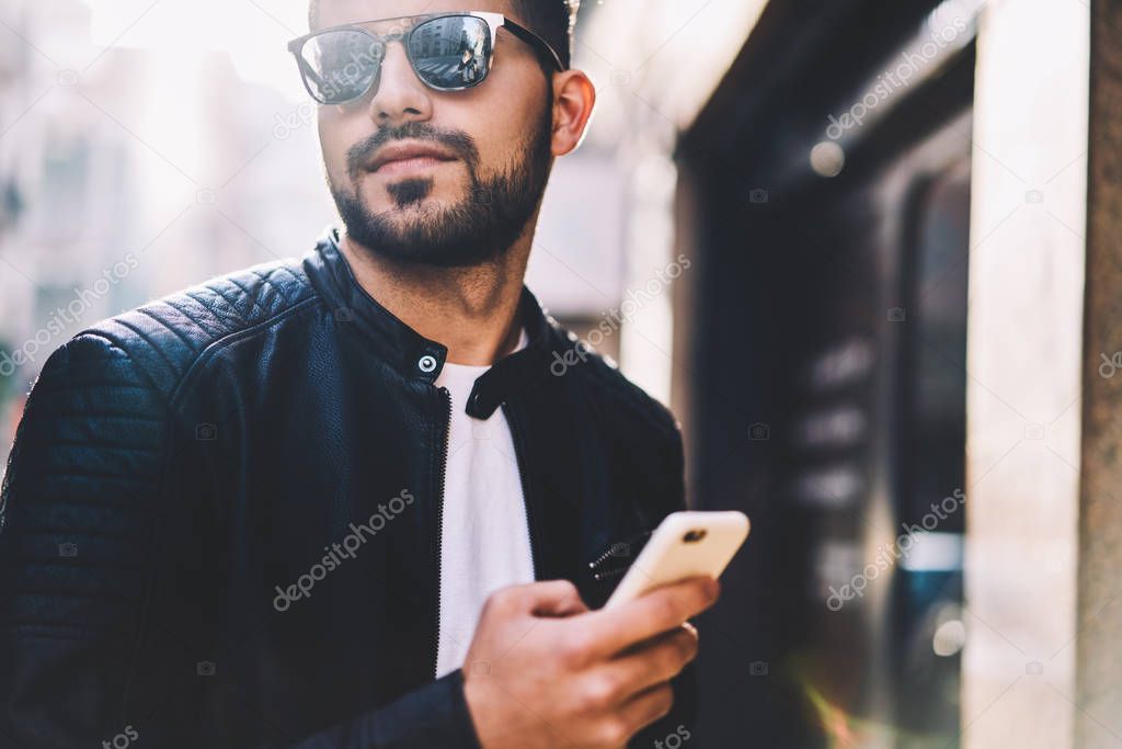Cropped image of bearded guy holding cellular share multimedia in networks looking away on street,handsome male sending text messages using application on smartphone for chatting online outdoors