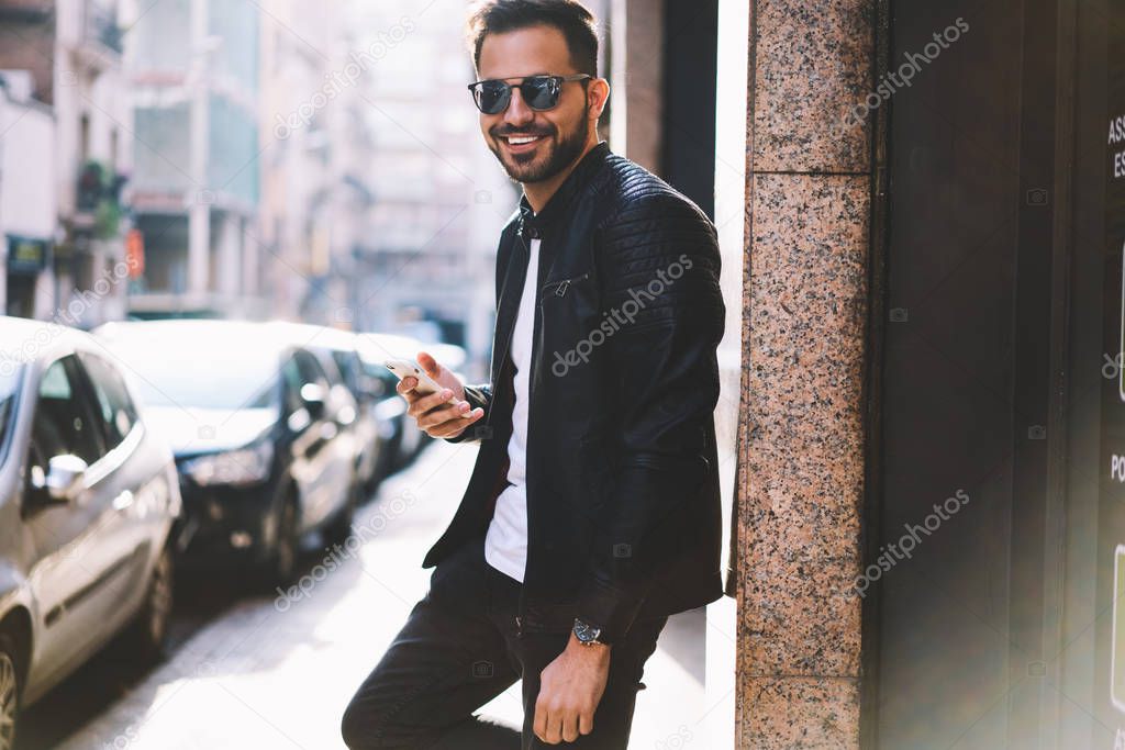 Half length portrait of cheerful hipster guy using smartphone standing on city street blogging in social networks,happy handsome male looking at camera chatting with friend online via telephone