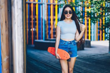 Hobi yaz boş zaman geçirmek için elinde kaykay tutarken kameraya gülümseyen güzel esmer saçlı esmer çekici genç kadın portresi. Teen açık havada duran güneş gözlüğü