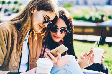Şık kadın blogcular kendi web sitelerinde yeni mesaj yayımlama ve açık havada elinde lezzetli kahve ile yeşil çimenlerin üzerinde oturan modern smartphone cihazlarda altındaki yorum okuma güneş gözlüğü