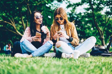 İki hippi kız lezzetli kahve içme ve güneşli havanın tadını çimenlerin üzerinde otururken telefonlar üzerinde sohbet online şık güneş gözlüğü. En yakın arkadaşlarım ile birlikte vakit cellulars