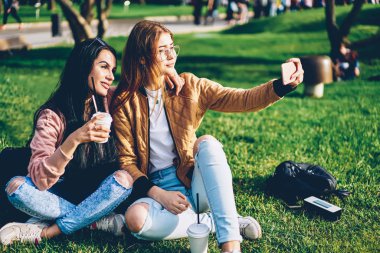 Parkta birlikte vakit smartphone kamera selfie yapmak için poz neşeli arkadaşlarım. Mutlu hippi kız trendy kıyafeti için hareket eden aygıt yolu ile sosyal ağlar uploading fotoğraf çekmek