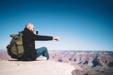 ABD, hipster adam mobil ağ iletişim arkadaş hiking kapsama ile memnun gezisi sırasında rock cliff üzerinde oturan görüntülü görüşme yapmak için iyi bir veri bağlantısı kullanarak sırt çantası ile erkek Gezgin  