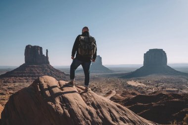 Monument Valley, vahşi çevre keşfetmek hedefe getting erkek yolcu Yolculuk tutkusu kuru çöl toprakları yüksek güzel sahne görünümünde bakarak kayada duran sırt çantası ile arkadan görünüşü adam