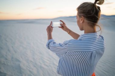 Genç kadın turist beyaz kum tepeleri, keşfetmek smartphone kamera Hipster kız Yolculuk tutkusu günbatımı sahne görünümünü tatil zevk cep telefonu kamera fotoğraf çekimi selfie yapma arkadan görünüm