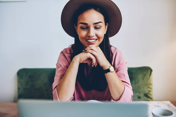 女性フリーランスの仕事をネットブック カフェのインテリアで過ごす時間にニュースを読んで満足肯定的な自由な時間の中にラップトップ コンピューターを使用してオンライン映画を見てスタイリッシュな帽子の若い女性を笑顔 — ストック写真