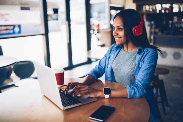 Cheerful female black model resting in coffee shop listening favorite music in headphones and chatting in networks.Beautiful dark skinned freelancer enjoying remote job typing publication on laptop