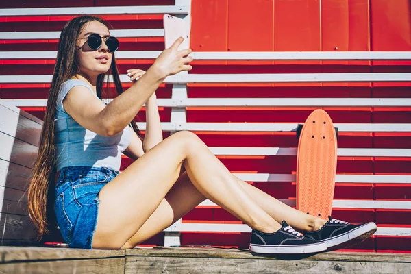 魅力的なカジュアル服を着た現代のスマート フォン デバイス休んで屋外スケート ボードでのフロント カメラで Selfie 写真を作るスタイリッシュなサングラス内気な少女 若い女性の携帯に写真を撮る — ストック写真