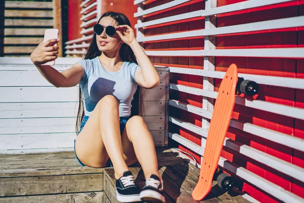 迷人的时髦的女孩在时尚的太阳镜享受夏天的天气和拍照前相机的现代智能手机发表在自己的博客更多的追随者坐在户外 — 图库照片