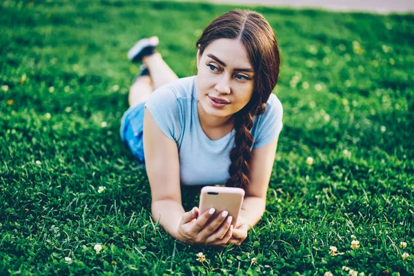 在浏览互联网网站时 在绿色草坪上 用手拿着电话的漂亮年轻女子在沉思 迷人的时髦女孩与辫子休息在草地上 而检查邮件的智能手机 — 图库照片