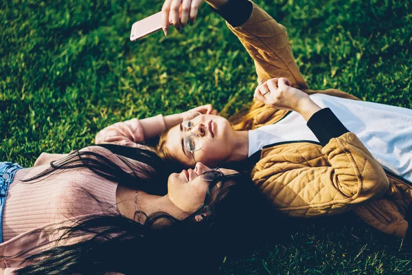 Two best friends bloggers in stylish sunglasses lying on green grass in park and making photos on front camera of modern smartphone device for own website during sunny weather in recreation time