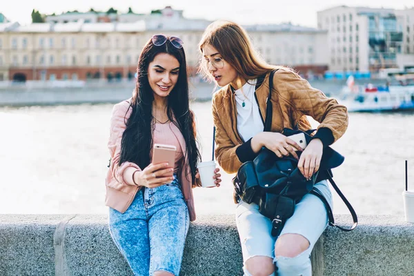 两个开朗最好的朋友与时尚的黑色背包和美味的咖啡手制作自拍图片通过新的照片应用安装在现代电话坐在城市宣传背景 — 图库照片