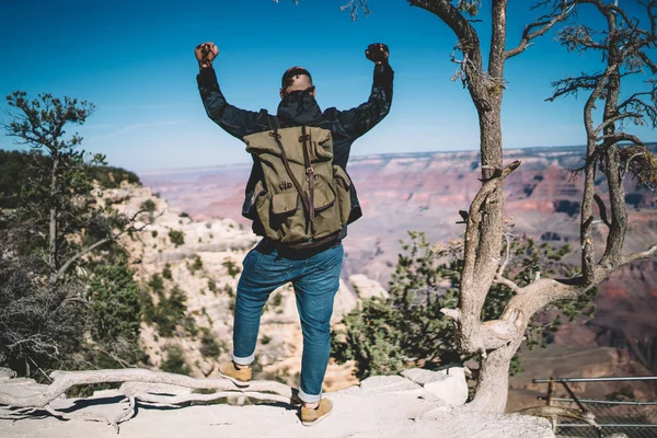 Back view of male tourist reaching top of mountain on hiking tour celebrating achievement rising hand,hipster guy with rucksack feeling freedom standing on rock peak enjoying exploring natur
