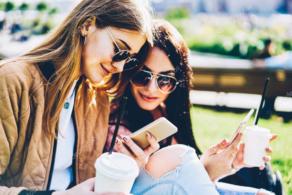 Stylish female bloggers in sunglasses publishing new posts on own websites and reading comments under it on modern smartphone devices sitting outdoors on green grass with tasty coffee in hands